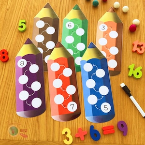 Può includere: Un set di sei carte di conteggio a forma di matita colorata con cerchi bianchi e frecce. Le carte sono numerate da 3 a 13. Le carte sono su una superficie di legno con numeri magnetici che indicano "3 + 6 = 9".