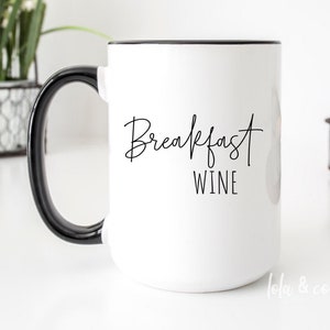 May include: White ceramic mug with a black handle and black rim. The mug has the text "Breakfast Wine" written in black cursive script.
