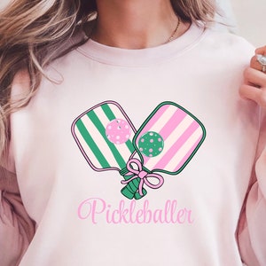 May include: Pink sweatshirt with a green and pink pickleball paddle design and the text "Pickleballer" in pink.