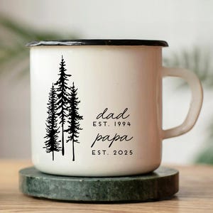 May include: White enamel mug with a black rim and handle. The mug features a black illustration of pine trees and the words "dad est. 1994 papa est. 2025" in black script. The mug sits on a green marble coaster.