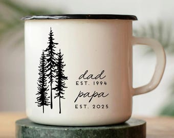 May include: White enamel mug with a black rim and handle. The mug features a black illustration of pine trees and the words "dad est. 1994 papa est. 2025" in black script. The mug sits on a green marble coaster.