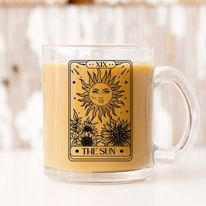 May include: A clear glass mug with a black and white illustration of the The Sun tarot card. The card features a sun with a face and sunflowers. The mug is filled with a light brown liquid.