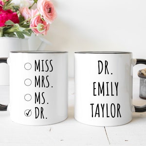 Puede incluir: Dos tazas de cer&aacute;mica blanca con asas y bordes negros. La primera taza presenta una lista con las opciones Miss, Mrs., Ms. y Dr., con una marca de verificaci&oacute;n junto a Dr. La segunda taza muestra el texto Dr. Emily Taylor en letras negras.