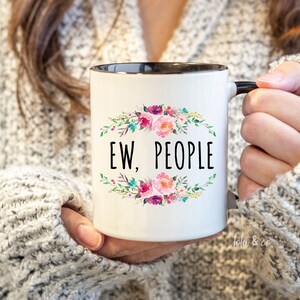 May include: White ceramic mug with a black rim and handle. The mug features a floral design with the text "EW, PEOPLE" in black.