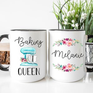 May include: Two white ceramic mugs with black handles. One mug has a blue and white illustration of a stand mixer with flowers and the text "Baking Queen". The other mug has a floral wreath with the name "Melanie" in the center.