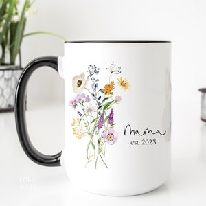 May include: White ceramic mug with a black handle and rim. The mug features a watercolor illustration of a bouquet of wildflowers in shades of purple, yellow, and orange. The text "Mama est. 2023" is printed in black cursive script below the flowers.