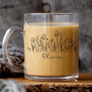 May include: A clear glass mug with a black and white illustration of mushrooms and grass. The mug is filled with a light brown liquid and the name "Olivia" is printed on the bottom of the design.