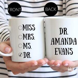 May include: Two white coffee mugs. The mug on the left has a checklist with the options "Miss", "Mrs.", "Ms.", and "Dr." with the last option checked. The mug on the right says "Dr. Amanda Evans".