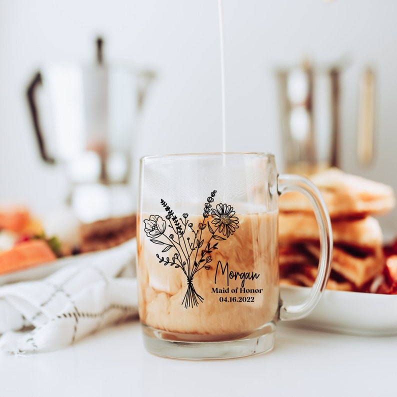 K&ouml;nnte beinhalten: Eine klare Glas-Tasse mit einem schwarzen Blumenmuster und dem Text "Morgan Maid of Honor 04.16.2022". Die Tasse ist mit Kaffee und Sahne gef&uuml;llt.