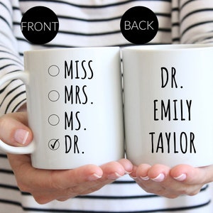 Puede incluir: Dos tazas de cer&aacute;mica blanca con texto en negro. La taza frontal muestra una lista de verificaci&oacute;n con las opciones MISS, MRS., MS. y DR., con una marca de verificaci&oacute;n junto a DR. La taza trasera muestra el texto DR. EMILY TAYLOR en negro.