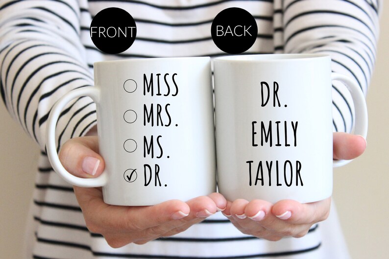 Puede incluir: Dos tazas de cer&aacute;mica blanca con texto en negro. La taza frontal muestra una lista de verificaci&oacute;n con las opciones MISS, MRS., MS. y DR., con una marca de verificaci&oacute;n junto a DR. La taza trasera muestra el texto DR. EMILY TAYLOR en negro.