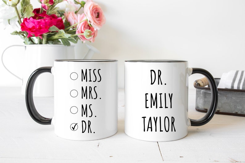 Puede incluir: Dos tazas de cer&aacute;mica blanca con asas y bordes negros. La primera taza presenta una lista con las opciones Miss, Mrs., Ms. y Dr., con una marca de verificaci&oacute;n junto a Dr. La segunda taza muestra el texto Dr. Emily Taylor en letras negras.