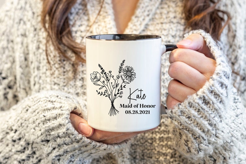 K&ouml;nnte beinhalten: Wei&szlig;e Keramiktasse mit schwarzem Rand und Henkel. Die Tasse zeigt eine schwarz-wei&szlig;e Illustration eines Blumenstrau&szlig;es und den Text "Kate Maid of Honor 08.28.2021".
