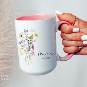 May include: White ceramic mug with a pink interior and handle. The mug features a floral design with a bouquet of watercolor flowers in shades of purple, yellow, and pink. The text "Mama est. 2023" is printed in black below the flowers.