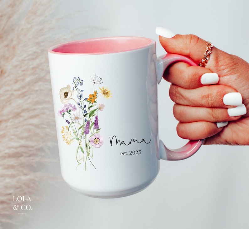 May include: White ceramic mug with a pink interior and handle. The mug features a floral design with a bouquet of watercolor flowers in shades of purple, yellow, and pink. The text "Mama est. 2023" is printed in black below the flowers.