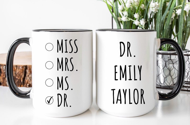 Puede incluir: Dos tazas de cer&aacute;mica blanca con asas y bordes negros. La taza de la izquierda presenta una lista con las opciones MISS, MRS., MS. y DR., con una marca de verificaci&oacute;n junto a DR. La taza de la derecha muestra el texto DR. EMILY TAYLOR en negro.