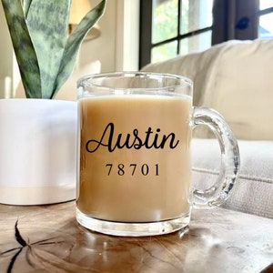 May include: A clear glass mug filled with a light brown beverage, featuring the word "Austin" in a cursive font and the number "78701" below it. The mug has a clear handle and sits on a wooden surface.