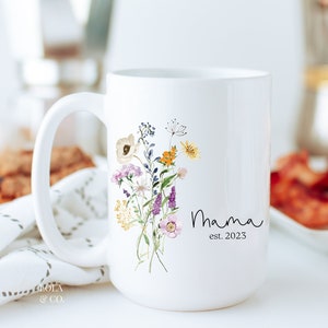 May include: White ceramic mug with a floral design and the text "Mama est. 2023".