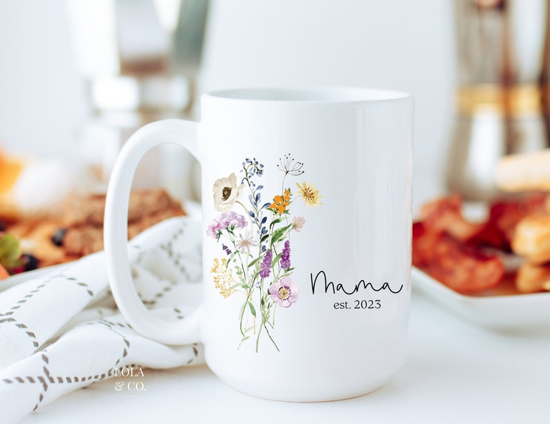 May include: White ceramic mug with a floral design and the text "Mama est. 2023".