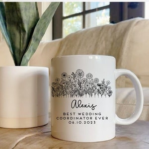 Może przedstawiać: Biały kubek ceramiczny z czarnym wzorem kwiatowym i tekstem "Alexis Best Wedding Coordinator Ever 06.10.2023".