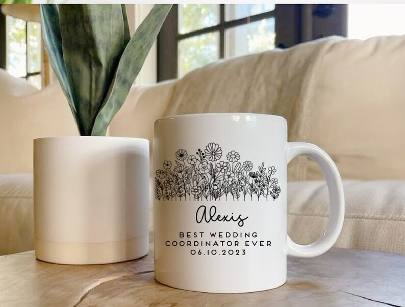 Może przedstawiać: Biały kubek ceramiczny z czarnym wzorem kwiatowym i tekstem "Alexis Best Wedding Coordinator Ever 06.10.2023".