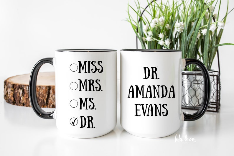 May include: Two white coffee mugs with black rims. One mug has a checklist with the options "Miss", "Mrs.", "Ms.", and "Dr." with the "Dr." option checked. The other mug has the text "Dr. Amanda Evans".