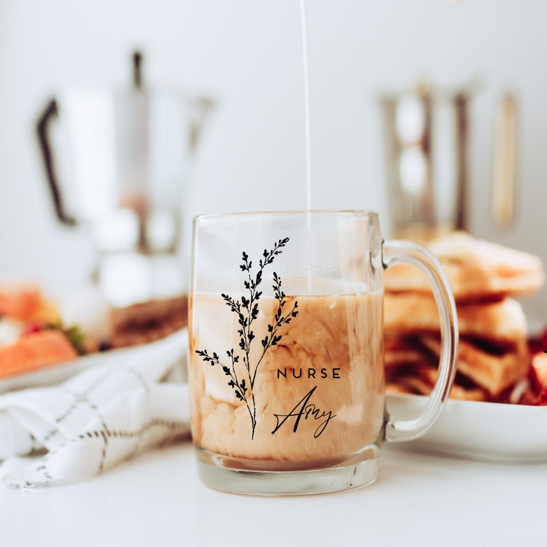 Pu&ograve; includere: Una tazza di vetro trasparente con un motivo floreale nero e il testo "NURSE Amy". La tazza &egrave; piena di caff&egrave; e panna.