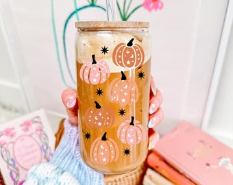 May include: A glass tumbler with a bamboo lid and a glass straw. The tumbler is decorated with a pattern of pink and orange pumpkins with stars and moons. The tumbler is filled with a brown liquid.
