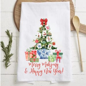 May include: White kitchen towel with a Christmas tree design made of Mahjong tiles, ornaments, and a red bow. The towel features the text "Merry Mahjong & Happy New Year!" in red script, with wrapped gifts at the base.