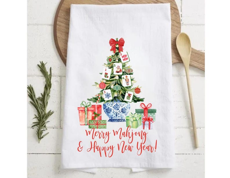 May include: White kitchen towel with a Christmas tree design made of Mahjong tiles, ornaments, and a red bow. The towel features the text "Merry Mahjong & Happy New Year!" in red script, with wrapped gifts at the base.