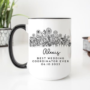 Może przedstawiać: Biały kubek ceramiczny z czarnym uchwytem. Kubek ma czarny zarys pola kwiat&oacute;w i tekst "Alexis Best Wedding Coordinator Ever 06.10.2023".