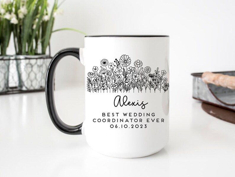 Może przedstawiać: Biały kubek ceramiczny z czarnym uchwytem. Kubek ma czarny zarys pola kwiat&oacute;w i tekst "Alexis Best Wedding Coordinator Ever 06.10.2023".