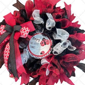 May include: A red, black, and white wreath with a sign that reads "Enjoy the little things". The sign has a ladybug on it. The wreath is decorated with bows and mesh.