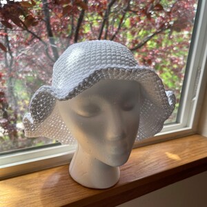 May include: White crochet bucket hat with a wide brim and a slightly ruffled edge. The hat is on a white mannequin head.