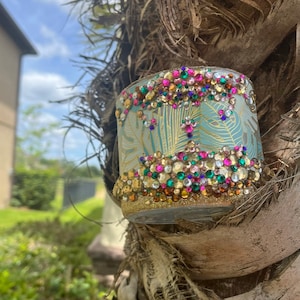 May include: A teal glass candle holder with a gold leaf design and a border of colorful rhinestones. The holder is attached to a tree trunk.