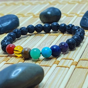 Peut inclure: Un bracelet en pierre de lave noire avec sept perles de chakra. Les perles sont rouge, orange, jaune, verte, bleue, indigo et violette. Le bracelet est sur un tapis en bambou tissé.