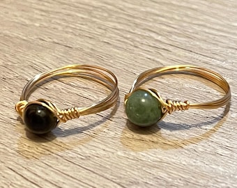 Puede incluir: Dos anillos hechos a mano con alambre. Un anillo presenta una piedra preciosa negra, el otro una piedra preciosa verde. Los anillos tienen una banda de alambre de color dorado y están sobre una superficie de madera.