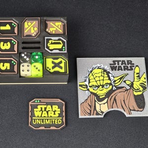 Könnte beinhalten: Ein schwarzes und grünes Würfeltablett mit einem Star Wars Unlimited Logo. Das Tablett hat sechs Fächer, jedes mit einem anderen Symbol. Es befinden sich vier Würfel im Tablett, zwei grüne, ein weißer und ein schwarzer. Eine graue Karte mit einem Star Wars Logo und einem Bild von Yoda befindet sich im Hintergrund.