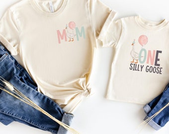May include: Two beige t-shirts with a goose design. The larger shirt says "MOM" with a goose holding a pink balloon. The smaller shirt says "ONE SILLY GOOSE" with a goose holding a pink balloon.