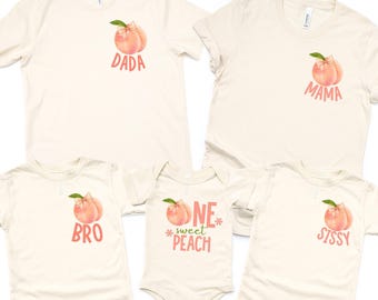 Op de afbeelding: Een verzameling crèmekleurige t-shirts en een romper, elk met een perzikafbeelding met een roze strik en groen blad. De shirts zijn voorzien van de tekst "DADA", "MAMA", "BRO" en "SISSY", terwijl de romper "ONE sweet PEACH" zegt.