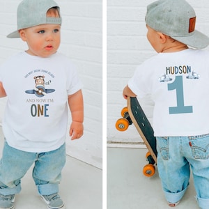 May include: A white t-shirt with a cartoon airplane design and the text "OH MY! HOW TIME FLIES! AND NOW I'M ONE." The back of the shirt has the name "HUDSON" and the number "1". The child is wearing blue jeans and a gray baseball cap.