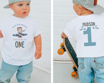 May include: A white t-shirt with a cartoon airplane design and the text "OH MY! HOW TIME FLIES! AND NOW I'M ONE." The back of the shirt has the name "HUDSON" and the number "1". The child is wearing blue jeans and a gray baseball cap.