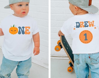 May include: A white t-shirt with a pumpkin graphic and the word "ONE" in blue and orange. The back of the shirt has the name "DREW" and the number "1" in a circle. The child is wearing blue jeans and a gray baseball cap.