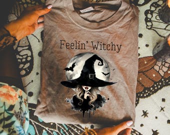 May include: A tan t-shirt with the text "Feelin' Witchy" above a graphic of a witch wearing a black hat in front of a full moon with bats. The witch is holding a finger to her lips. The shirt is folded.
