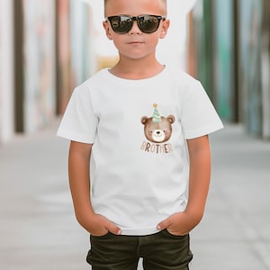 May include: White t-shirt with a brown bear wearing a blue and yellow party hat. The bear is looking to the right and the text "BROTHER" is printed below the bear.