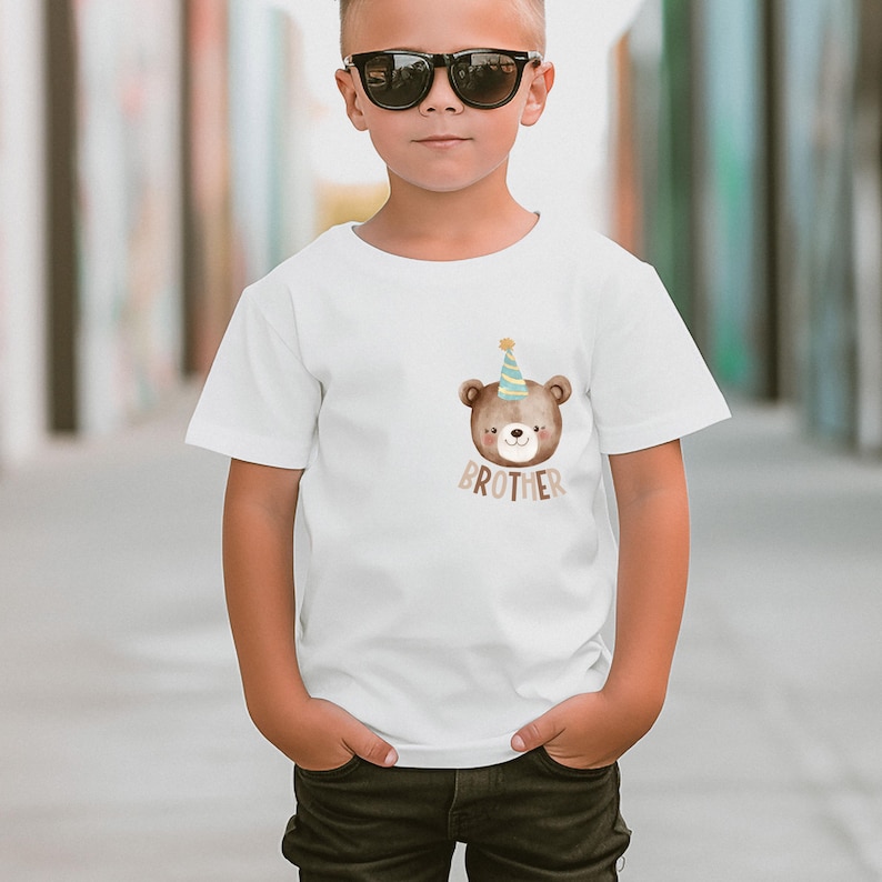 May include: White t-shirt with a brown bear wearing a blue and yellow party hat. The bear is looking to the right and the text "BROTHER" is printed below the bear.