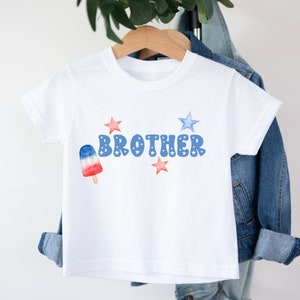K&ouml;nnte beinhalten: Wei&szlig;es T-Shirt mit einem blau-roten Eis am Stiel, dem Wort "BROTHER" in blauen Buchstaben und roten und blauen Sternen.