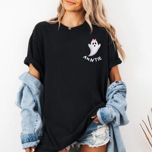 May include: Black t-shirt featuring a white ghost with a pink bow and the word "AUNTIE" in white script. The shirt has short sleeves and a relaxed fit. The model is wearing a denim jacket.