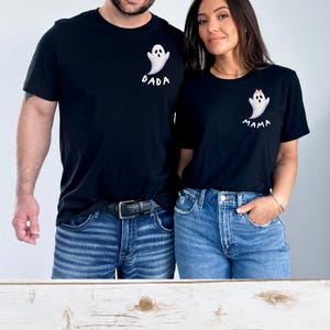 May include: Black t-shirts with matching ghost designs. One shirt says "DADA" and the other says "MAMA". The ghost designs are white with black eyes and a small pink bow on the "MAMA" shirt. Both shirts are paired with blue jeans.