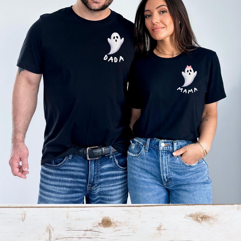 May include: Black t-shirts with matching ghost designs. One shirt says "DADA" and the other says "MAMA". The ghost designs are white with black eyes and a small pink bow on the "MAMA" shirt. Both shirts are paired with blue jeans.